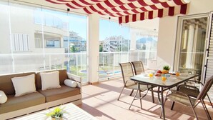 A balcony with a red and white striped awning, a sofa, a dining table with chairs, and a view of buildings and a pool. - Apartament Los Mares by DENIA COSTA (Dénia)