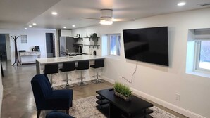 Un soggiorno moderno con una grande televisione, poltrone blu scuro, un tavolino da caffè e un'area cucina sullo sfondo.