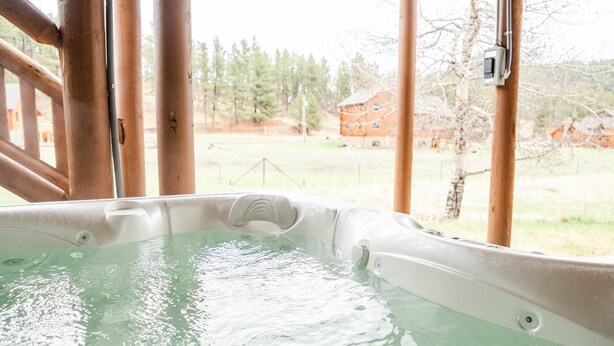 A hot tub with a view of a wooden cabin and trees in the background. - Welcome to Goldfinch! (Hill City)