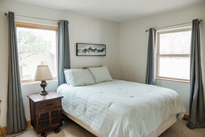 A bedroom with a bed, a nightstand with a lamp, a window with curtains, and a wall-mounted picture.