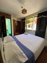 Habitación de hotel con una cama grande, ventilador de techo, ventana con cortinas y vistas al exterior.