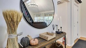 A hallway with a round mirror, a vase with dried plants, a wooden console table with candles and a woven basket.