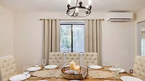 A dining room with a wooden table set for six, beige upholstered chairs, a chandelier, and a window with curtains.