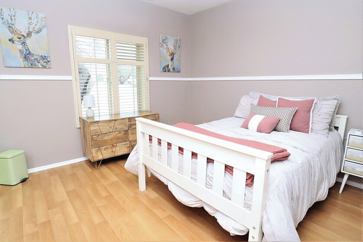 A bedroom with a bed, a dresser, a window with blinds, and artwork on the walls.