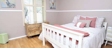 A bedroom with a bed, a dresser, a window with blinds, and artwork on the walls.