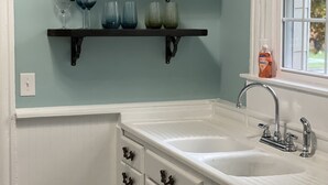 A kitchen with white cabinets, a double sink, and two shelves holding wine glasses. - Adorable 1899 historic “The Butter Bean Lady” storybook home (Columbus)