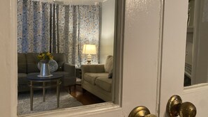 A room with a sofa, a small table with a lamp, and curtains with a floral pattern. - Adorable 1899 historic “The Butter Bean Lady” storybook home (Columbus)