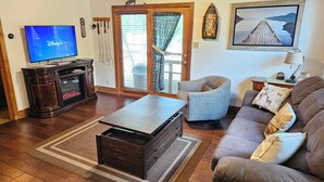 A living room with a TV, wooden entertainment unit, sofa, armchair, and a view of the outdoors through a glass door.