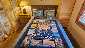 A bedroom with a bed featuring a 'Lake' themed quilt, a wooden headboard, a nightstand with a lamp, and a wall with a fish decoration.