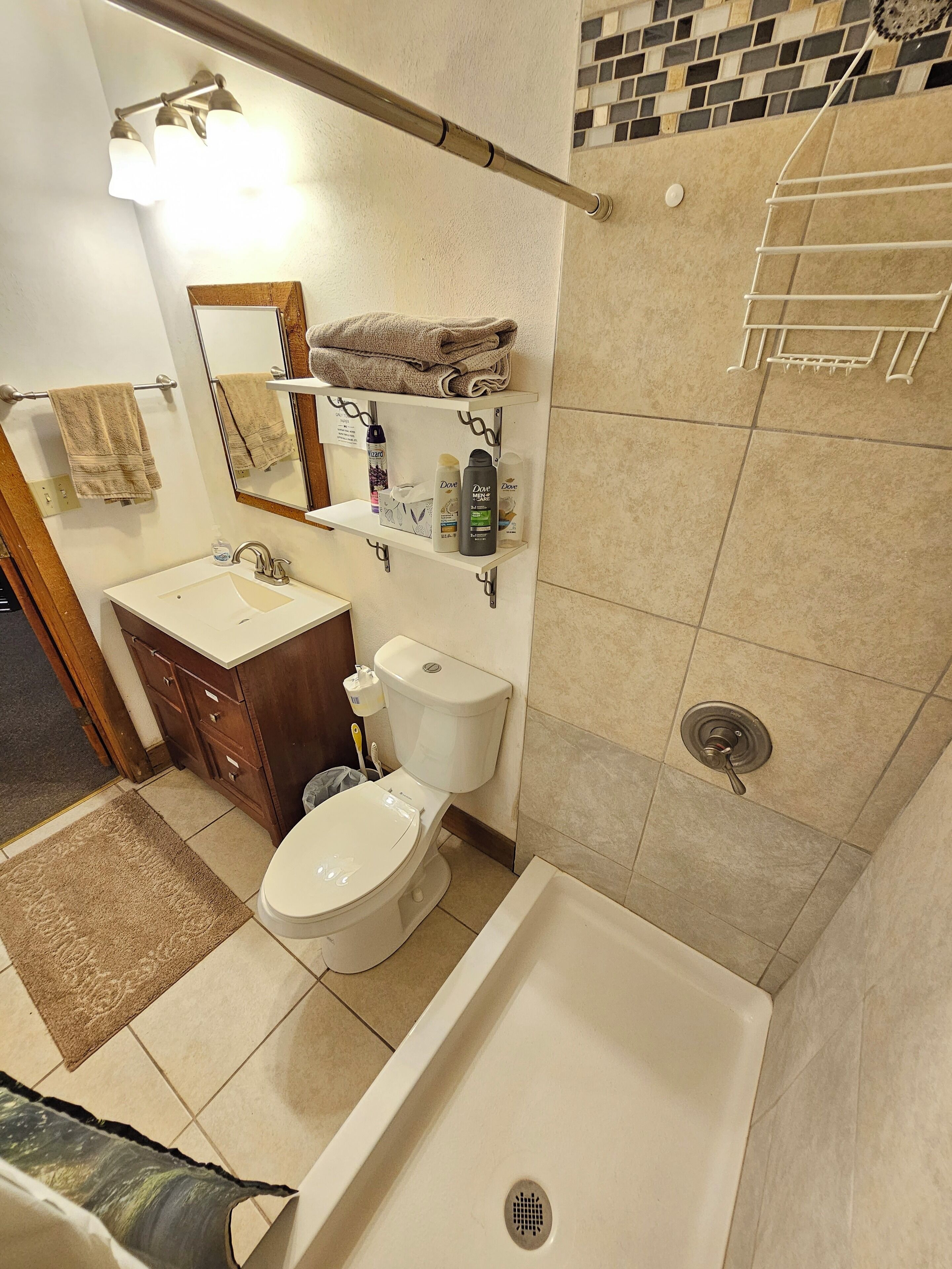 Une salle de bain comprenant des toilettes, un lavabo et une baignoire. On y trouve des serviettes suspendues à un porte-serviettes, une étagère avec des articles de toilette et un espace douche équipé d’un autre porte-serviettes.