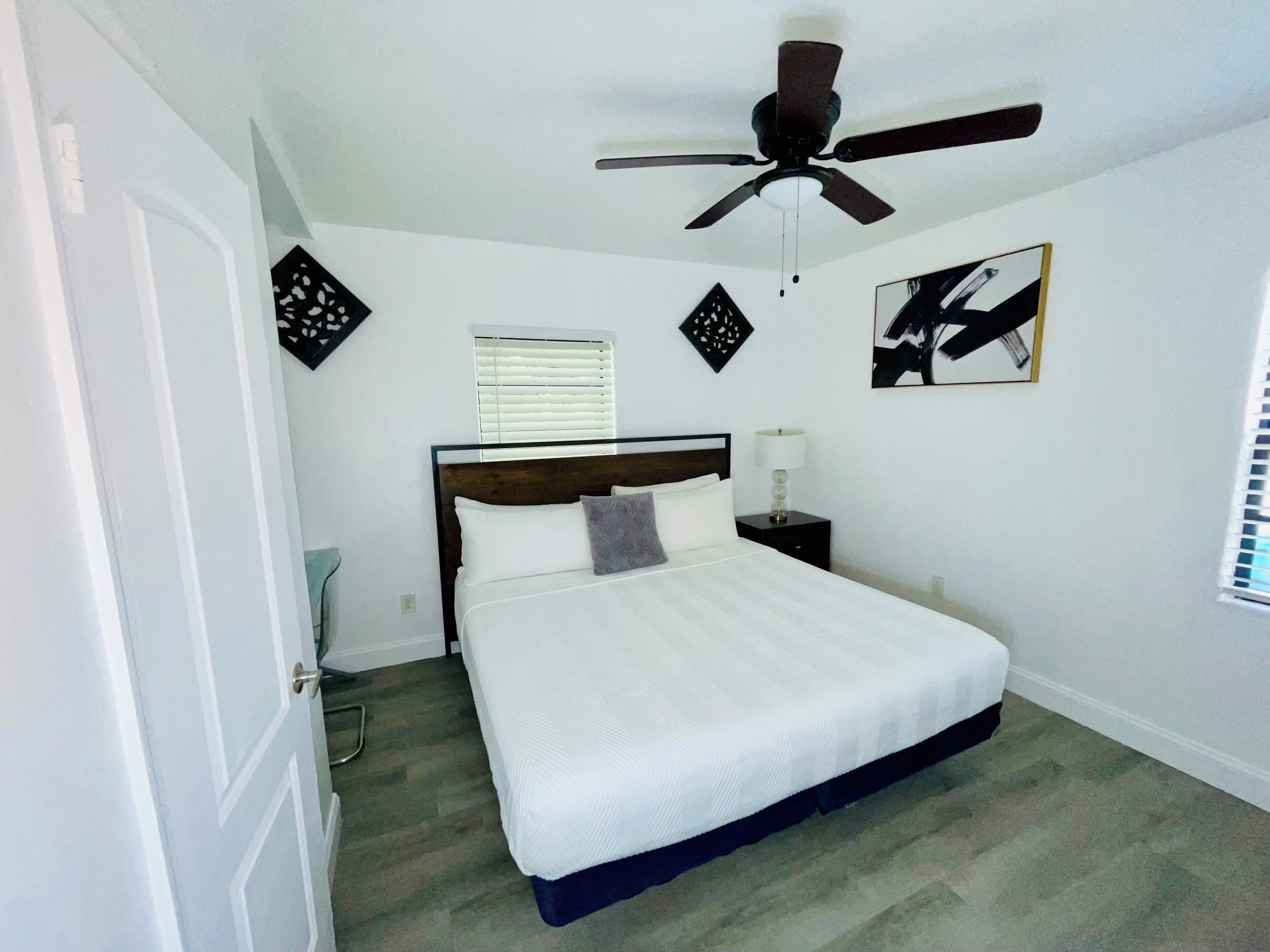 Ein Schlafzimmer mit Bett, Deckenventilator, Nachttisch, Lampe, Fenster mit Jalousien und einer Tür.