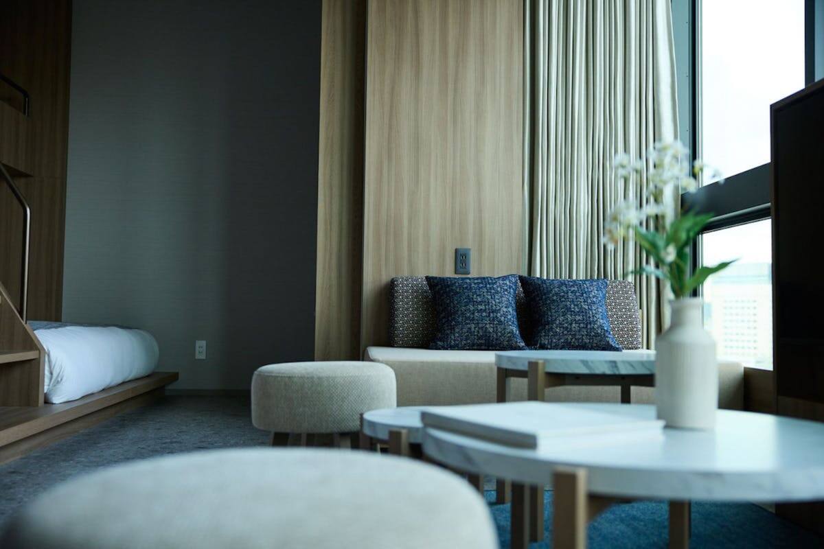 A modern hotel room with a bed, a sofa, two ottomans, a small table, and a large window with curtains.