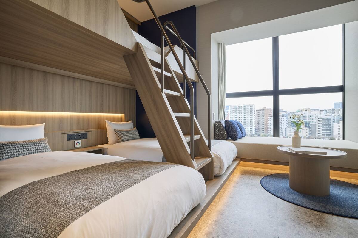 A modern hotel room with a large bed, a second bed, a staircase, a window with a city view, and a small table with a vase.