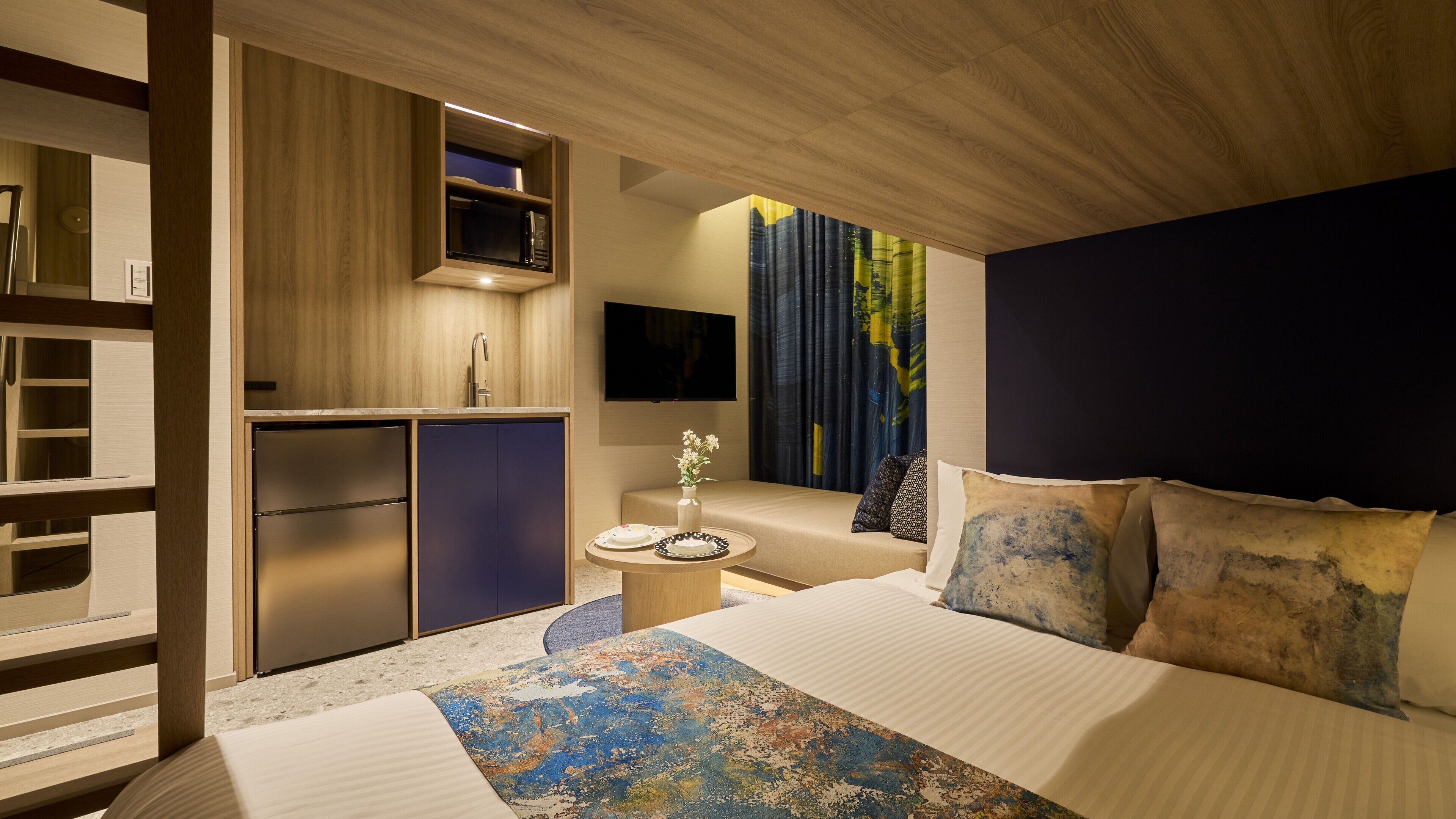 A modern hotel room with a bed, a small kitchenette, a flat-screen TV, and a view of the city.