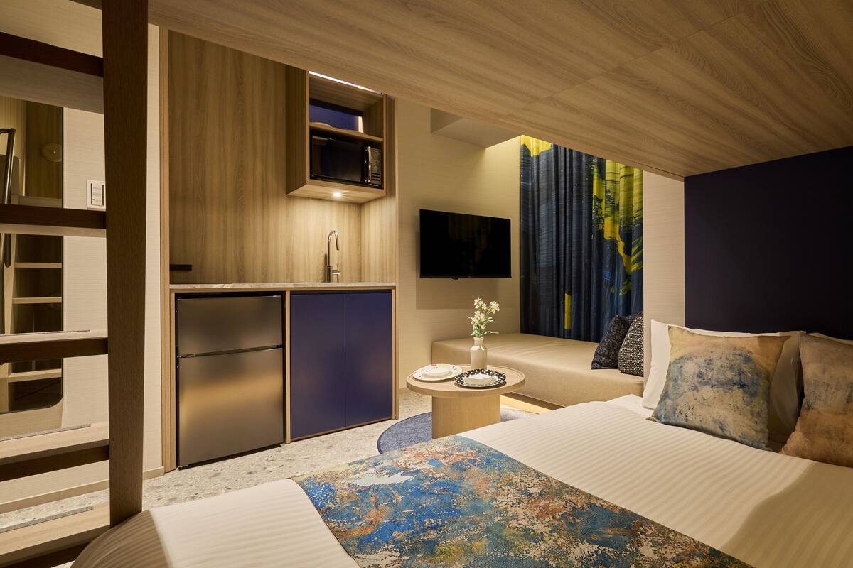 A modern hotel room with a bed, a small kitchenette, a flat-screen TV, and a view of the city.