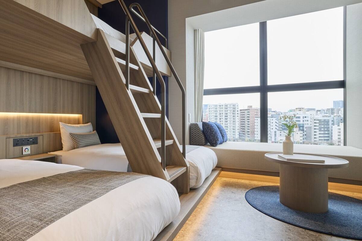 Une chambre d’hôtel moderne avec un grand lit, un deuxième lit plus petit, un escalier, une table de chevet, une commode, une fenêtre donnant sur la ville et une table basse ronde.