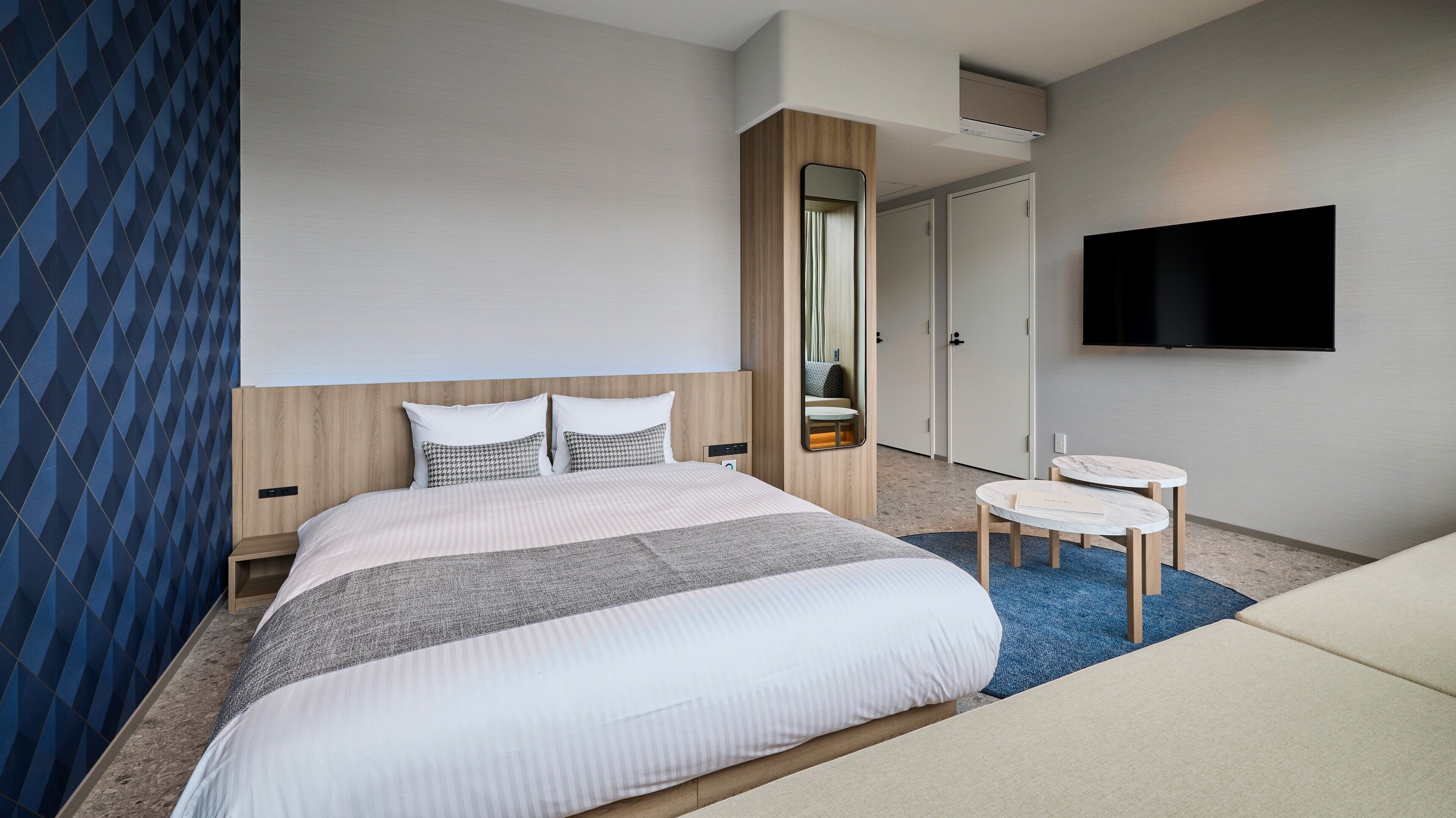 A modern hotel room with a large bed, a bedside table, a TV, and a small desk with a chair.