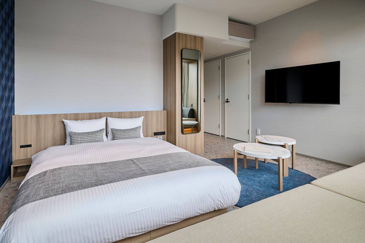 A modern hotel room with a large bed, a bedside table, a TV, and a small desk with a chair.