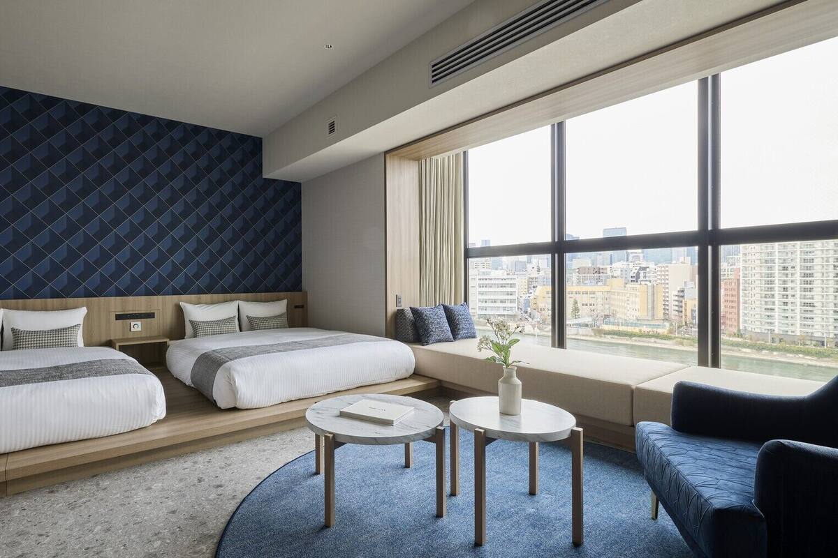 Une chambre d’hôtel avec deux lits, un canapé, une petite table et une grande fenêtre donnant sur la ville.