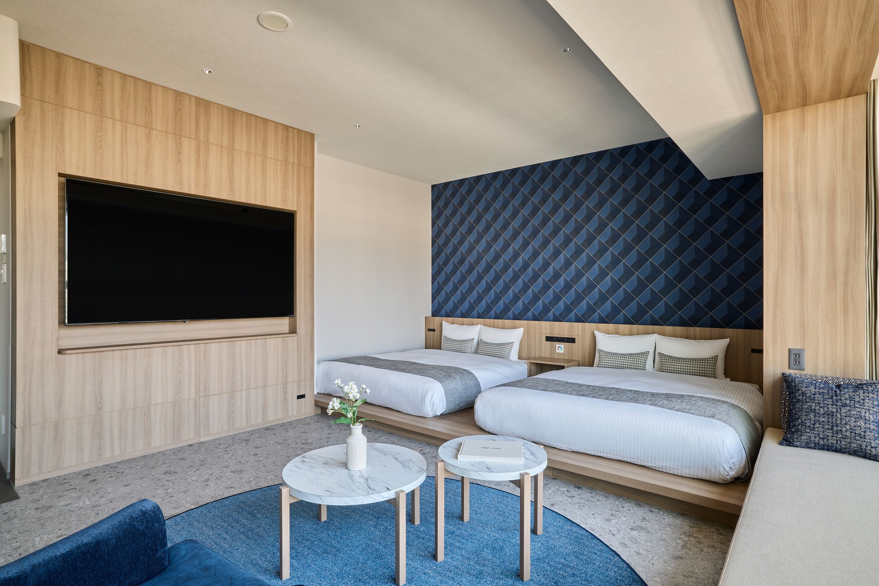 Habitación de hotel moderna con dos camas, un televisor de pantalla plana, una pequeña zona de estar con una mesa y sillas, y una alfombra azul.