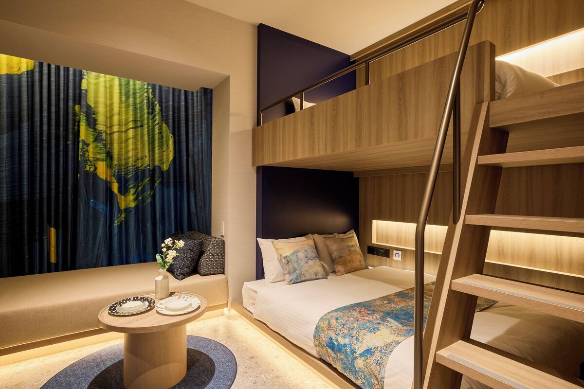 A modern hotel room with a bunk bed, a round table with plates, a wall art piece, and a bed with a patterned blanket.
