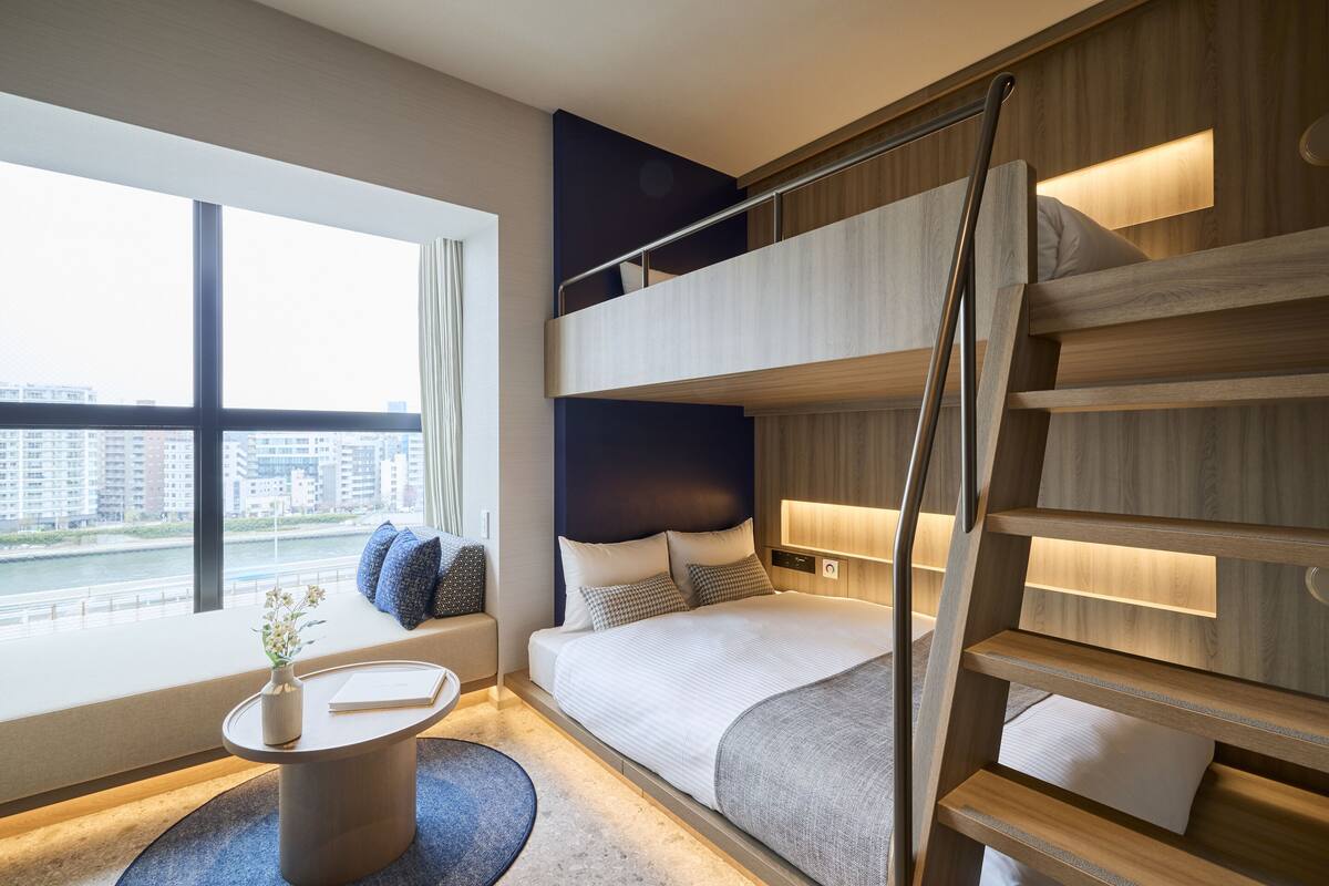 A modern hotel room with a large window, a bed with a headboard, a small table with a vase, and a staircase.