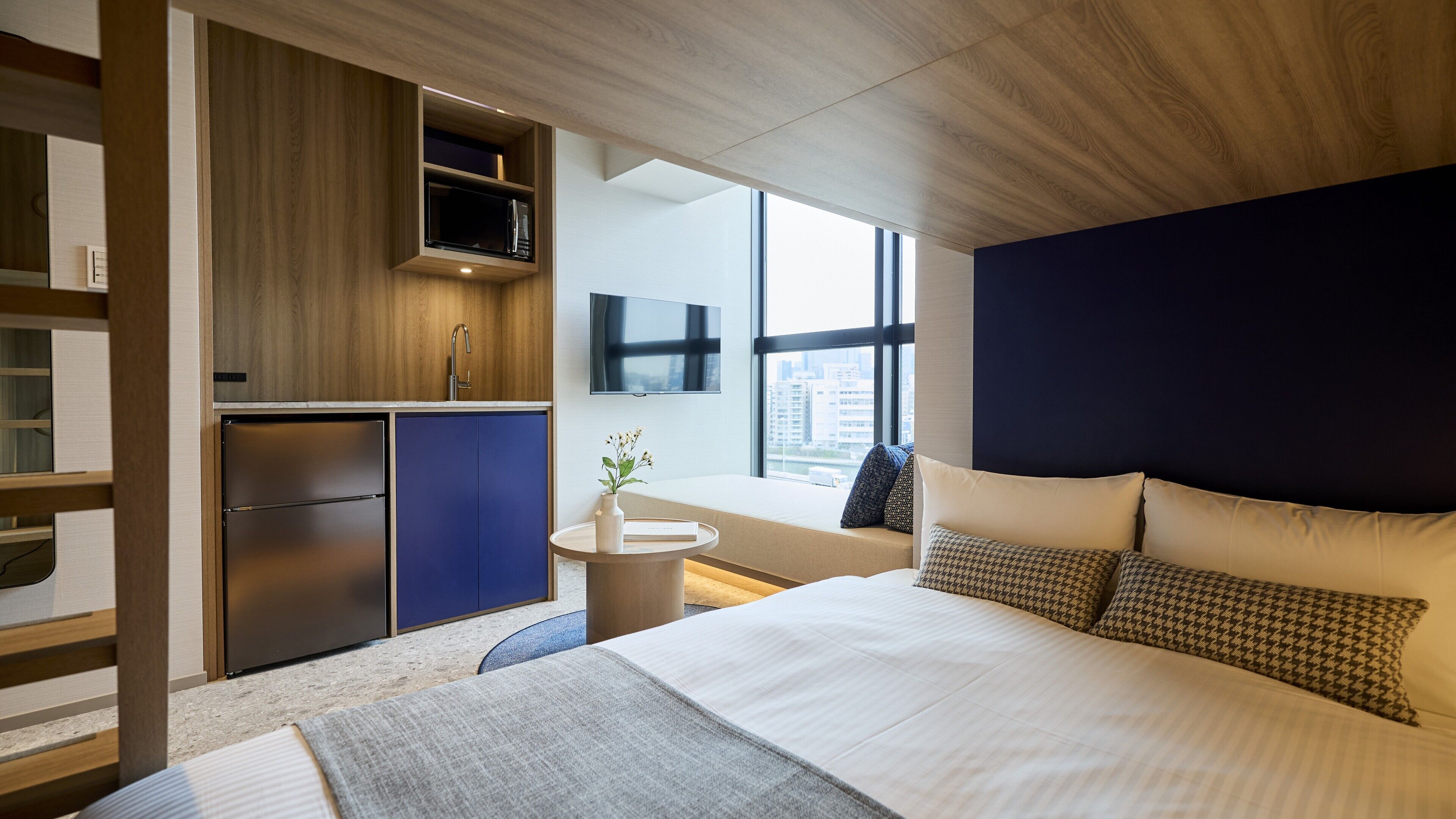 A modern hotel room with a bed, a built-in kitchen, a small round table, and a large window offering a city view.