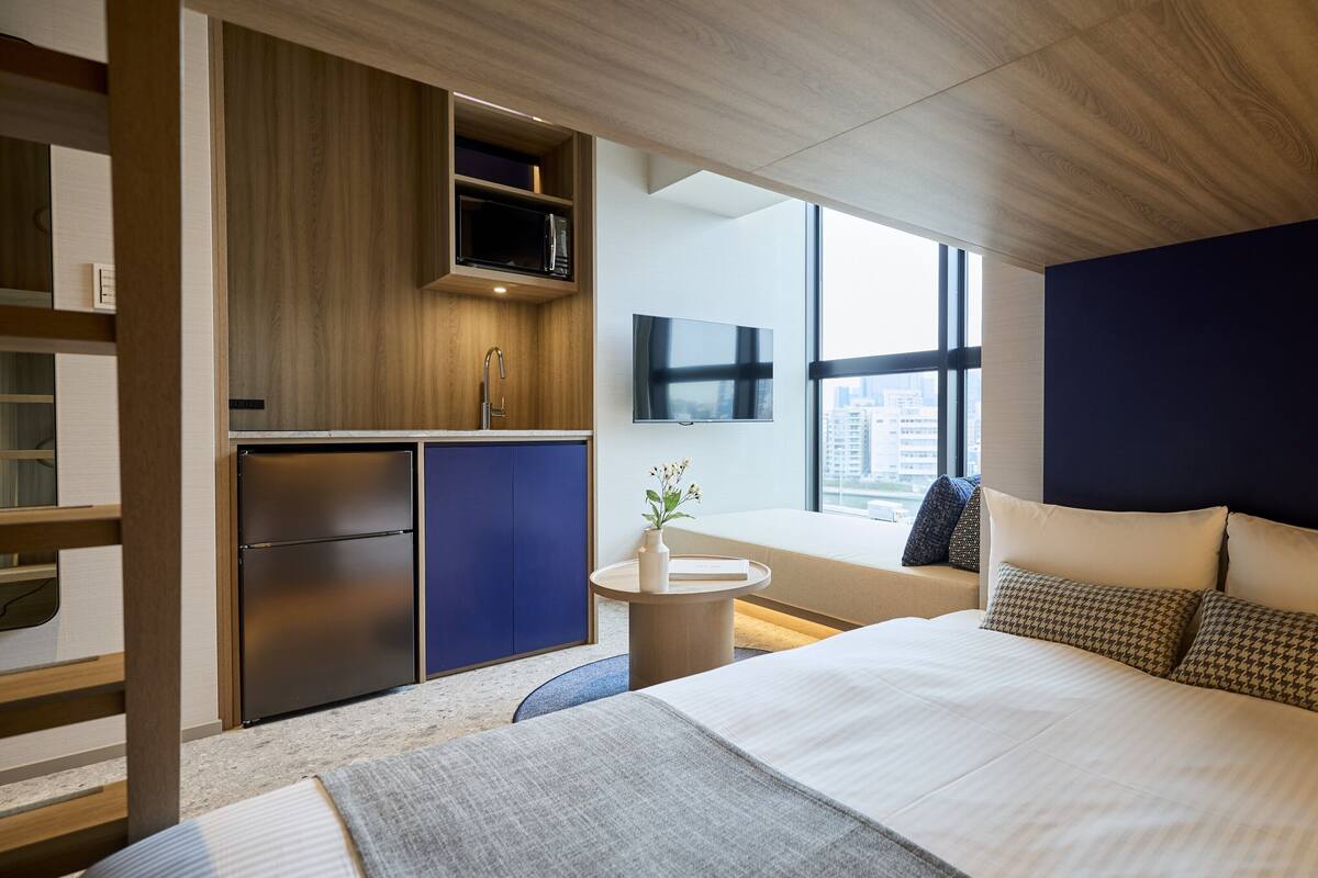 A modern hotel room with a bed, a built-in kitchen, a small round table, and a large window offering a city view.