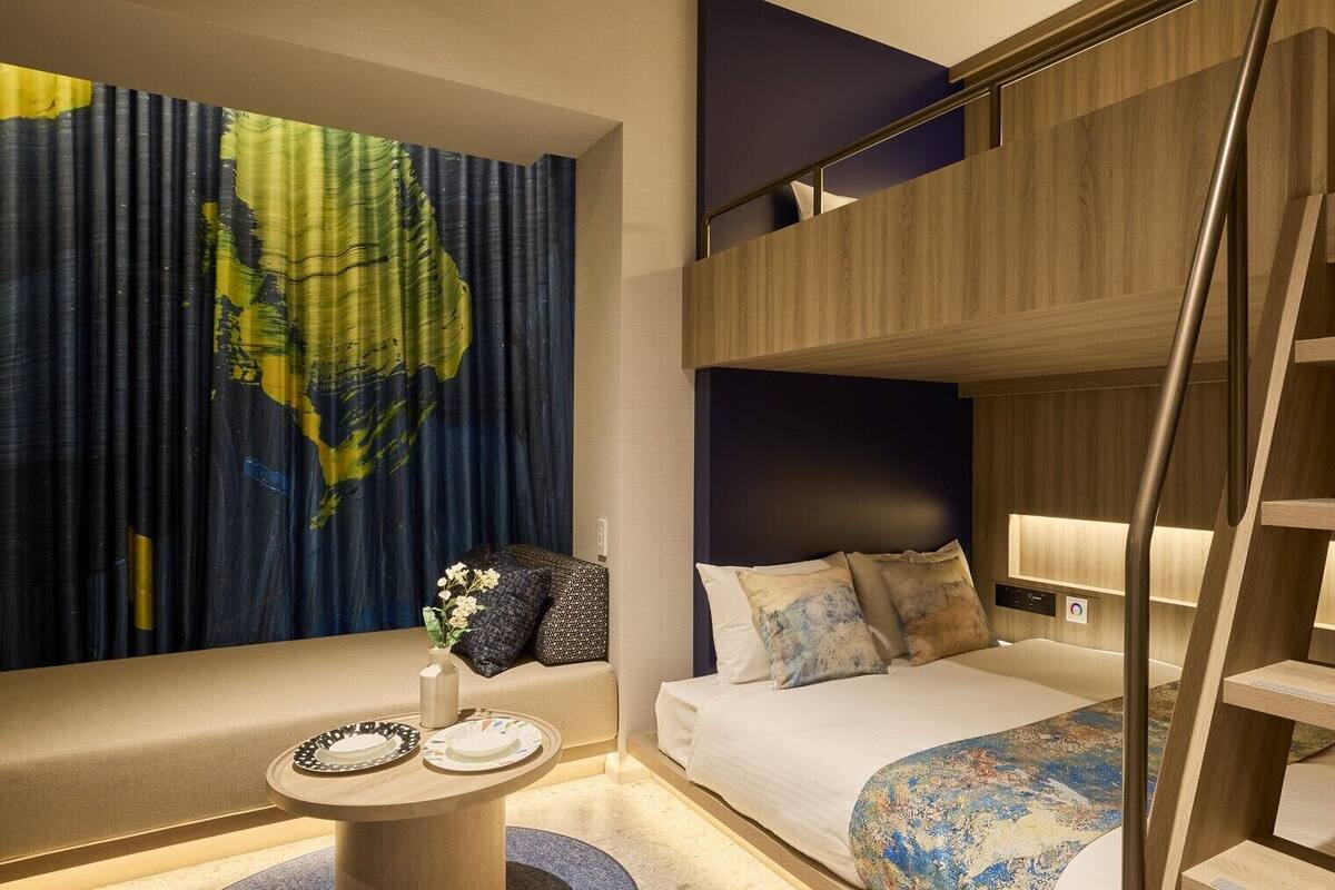 Une chambre d’hôtel moderne avec un lit superposé, une table ronde avec des assiettes, un vase décoratif et un grand tableau abstrait accroché au mur.