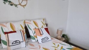 Un lit recouvert d’une couvre-lit à motifs floraux, plusieurs coussins aux motifs abstraits, un plaid jaune et une petite table ronde avec une lampe.