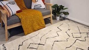 Un salon moderne avec un canapé gris, un plaid moutarde et des coussins à motifs. On y trouve deux étagères en bois avec des plantes suspendues et une petite plante en pot posée sur le sol.