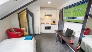 Una habitación de hotel compacta con dos camas, un escritorio con una computadora portátil, un televisor montado en la pared, una kitchenette y una silla roja.