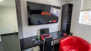 Et moderne værelse med et stort fladskærms-tv, der viser Netflix-logoet, en bærbar computer, en spillekonsol og en rød stol.