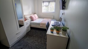 A bedroom with a bed, a desk with a laptop, a radiator, a window with blinds, and a wardrobe.