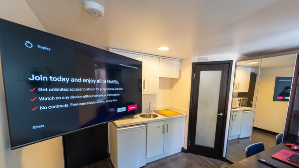 A modern kitchen with white cabinets, a black countertop, and a sink. A flat-screen TV mounted on the wall displays a Netflix sign. - Large studio 10 minute train journey to the city! (Birmingham)