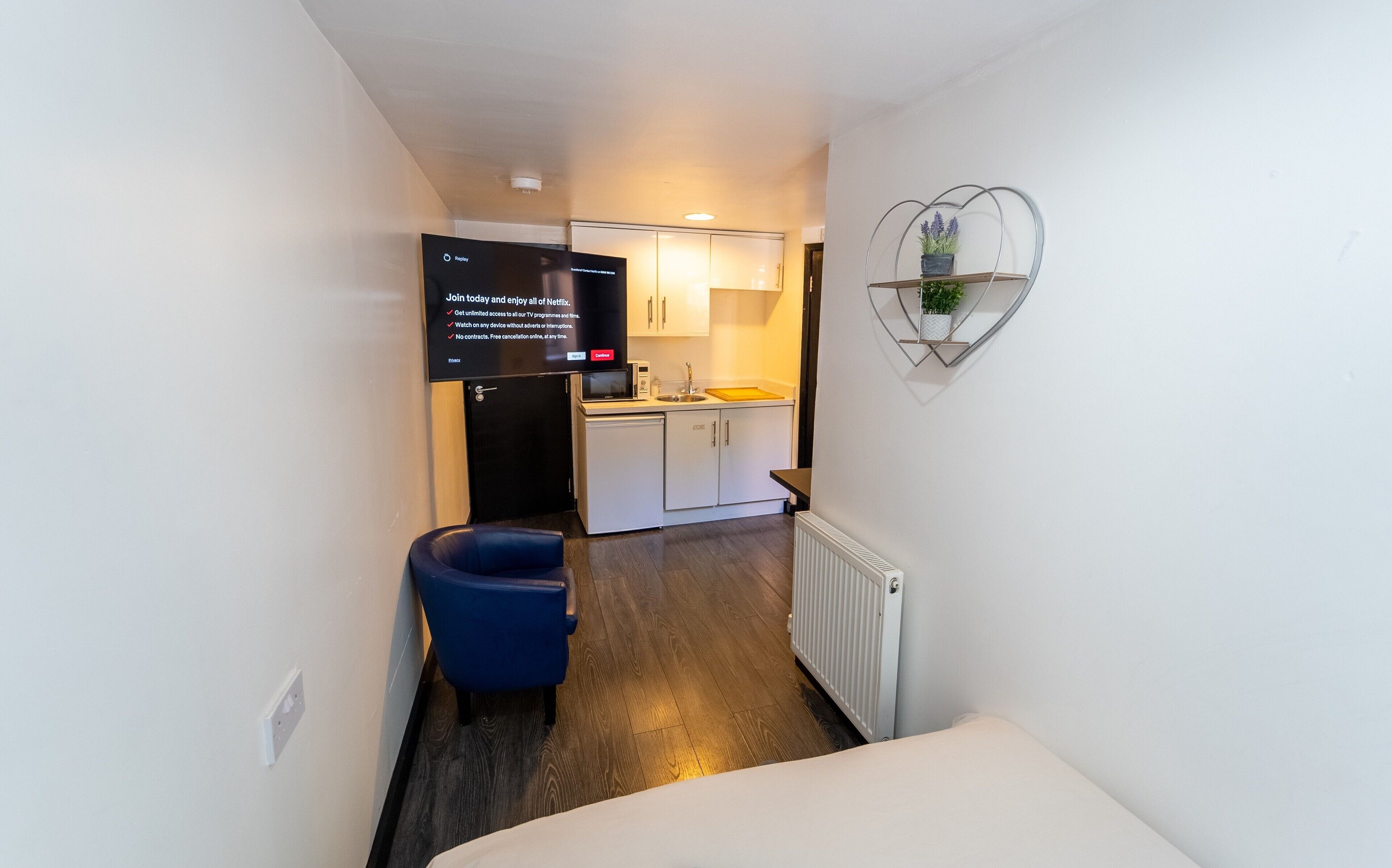 A narrow hallway leading to a kitchenette with a microwave, sink, and cabinets. A flat-screen TV is mounted on the wall displaying a Netflix sign. A blue chair is placed in the hallway, and a decorative wall piece hangs above the sink.
