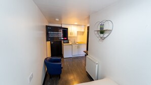 A narrow hallway leading to a kitchenette with a microwave, sink, and cabinets. A flat-screen TV is mounted on the wall displaying a Netflix sign. A blue chair is placed in the hallway, and a decorative wall piece hangs above the sink. - Large studio 10 minute train journey to the city! (Birmingham)