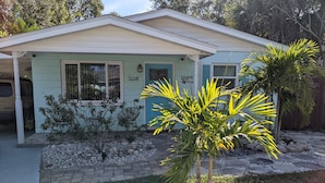 A single-story house with a light blue exterior, a front porch with two chairs, and a small garden area with palm trees and a gravel pathway. - "Happy Ours" Gulfport - charming home, walk to beach and town (Gulfport)