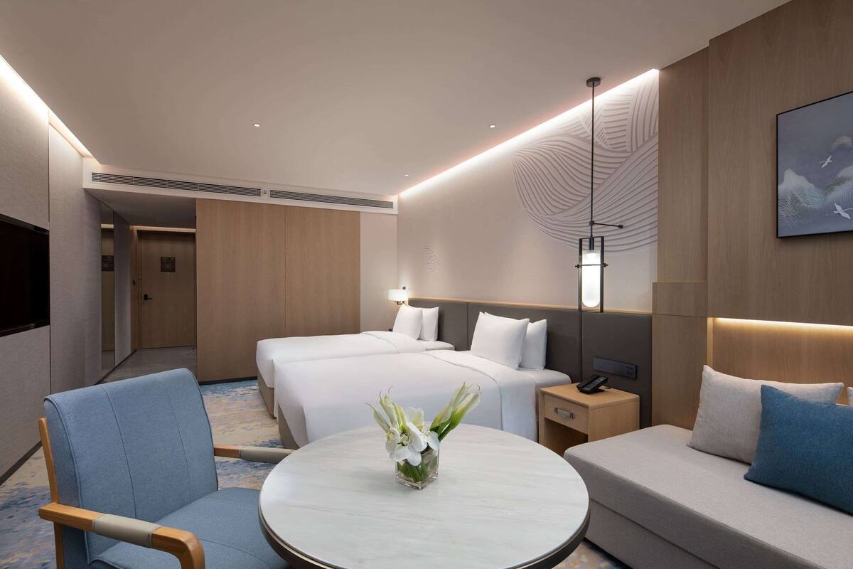 Quarto de hotel moderno com uma cama grande, um sofá, uma mesa redonda com um vaso de flores e uma TV montada na parede.