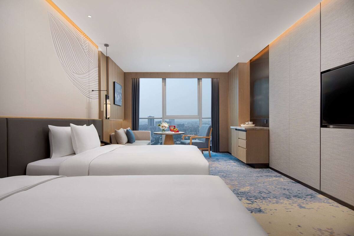 Quarto de hotel com duas camas, uma escrivaninha, uma cadeira, uma televisão e uma janela grande.