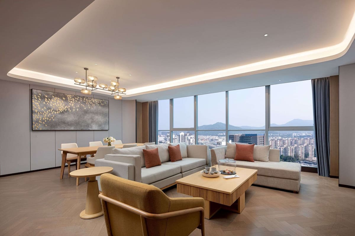 Uma sala de estar moderna com sofá, mesa de centro e cadeiras, grandes janelas com vista para a cidade e um lustre.