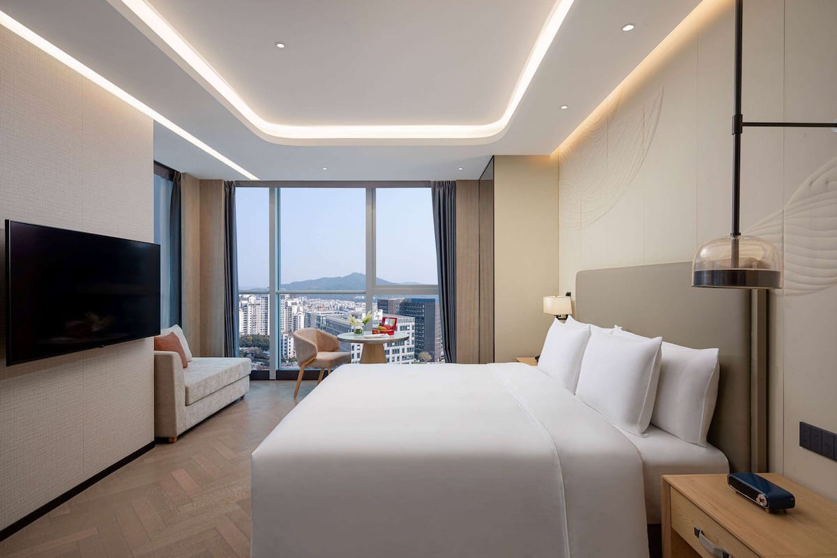 Quarto de hotel moderno com uma cama grande, televisão de tela plana, sofá, mesa redonda e vista para a cidade.