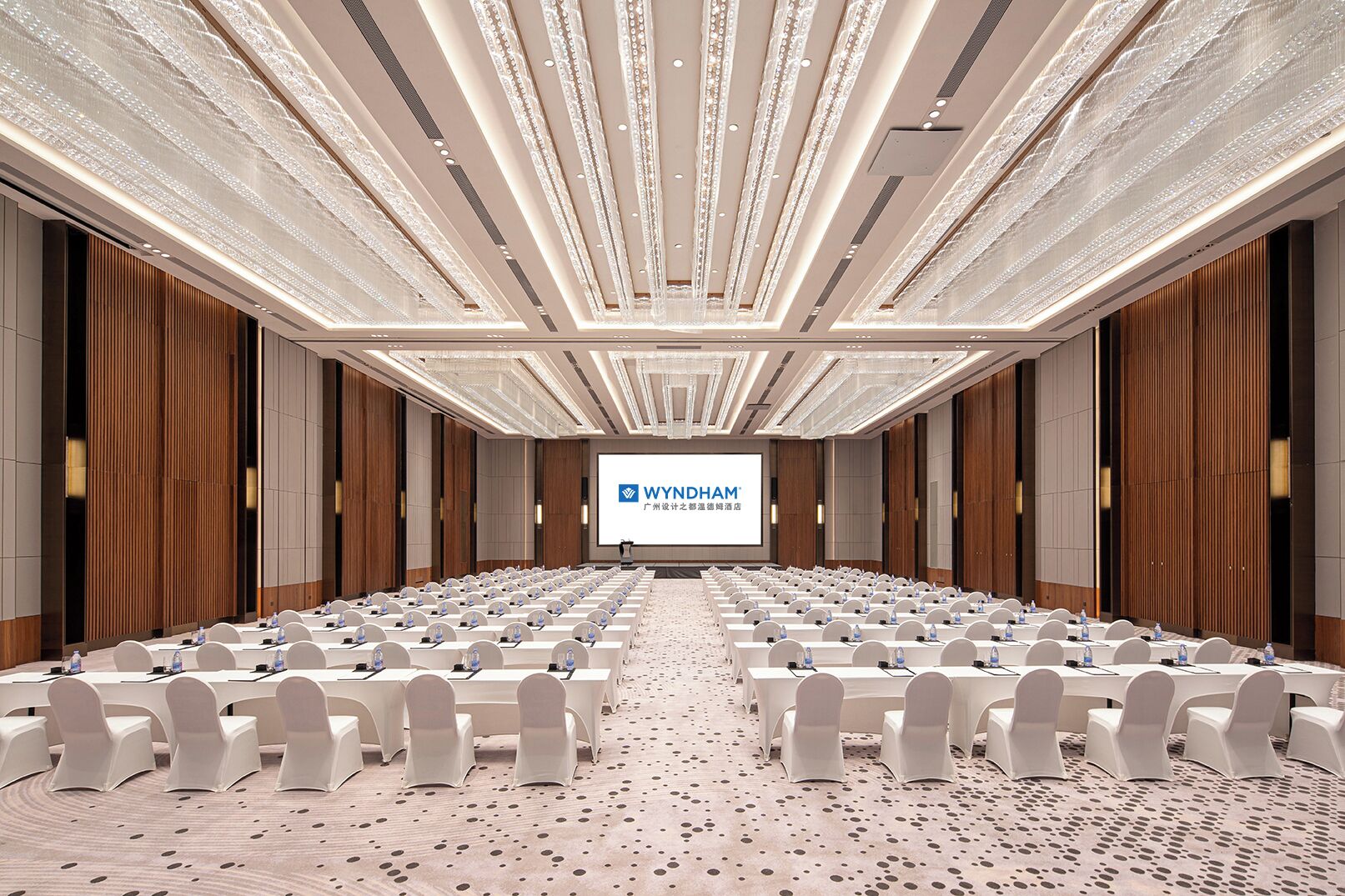 Una sala conferenze con file di tavoli e sedie, un grande schermo che mostra il logo WYNDHAM e una moquette fantasia.