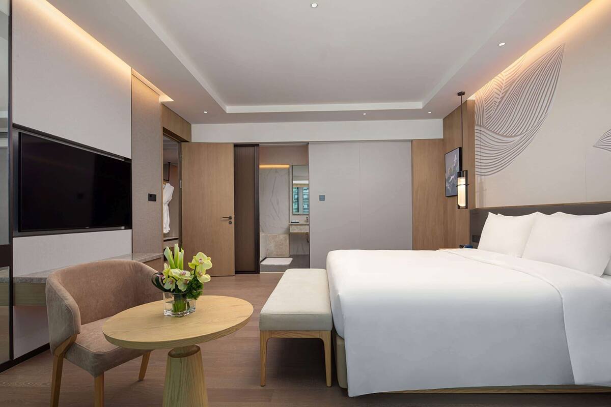 Quarto de hotel moderno com uma cama grande, televisão de tela plana, mesa redonda com um vaso de flores e um banco de madeira.