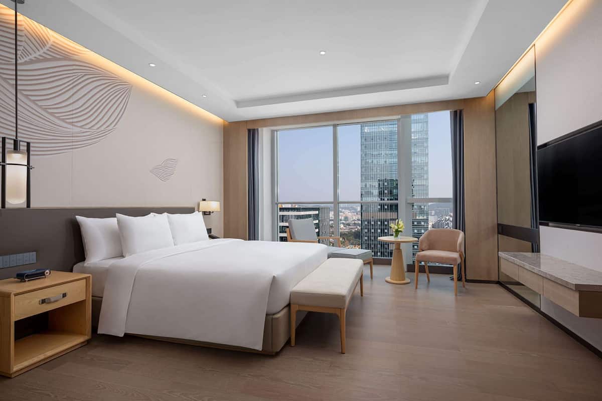 Quarto de hotel moderno com uma cama grande, uma escrivaninha, uma cadeira, uma televisão e varanda com vista para a cidade.