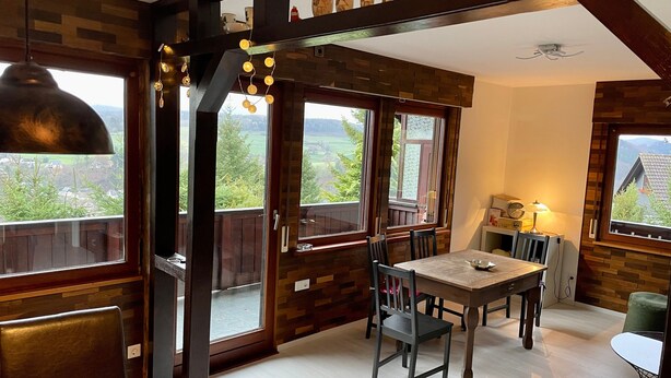 A dining area with a table and chairs, wooden beams, and large windows with a view of greenery. - Holiday Home 'Balke' with Mountain View, Garden, Terrace & Wi-Fi (Hatzfeld)