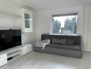 Un salon moderne doté d’un téléviseur à écran plat, d’un canapé gris, d’un meuble blanc et d’une fenêtre donnant sur des arbres.