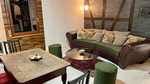 A living room with a brown leather sofa, a glass coffee table, a wooden dining table, and a green ottoman. - Holiday Home 'Balke' with Mountain View, Garden, Terrace & Wi-Fi (Hatzfeld)