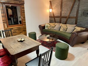 A living room with a brown leather sofa, a glass coffee table, a wooden dining table, and a green ottoman. - Holiday Home 'Balke' with Mountain View, Garden, Terrace & Wi-Fi (Hatzfeld)