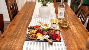 A wooden dining table with a variety of cheeses, meats, and fruits, accompanied by a bottle of wine and glasses. - Wine Trail Retreat w/ Fire Pit, Cold Plunge, Grill, Walk to Downtown JC! (Johnson City)